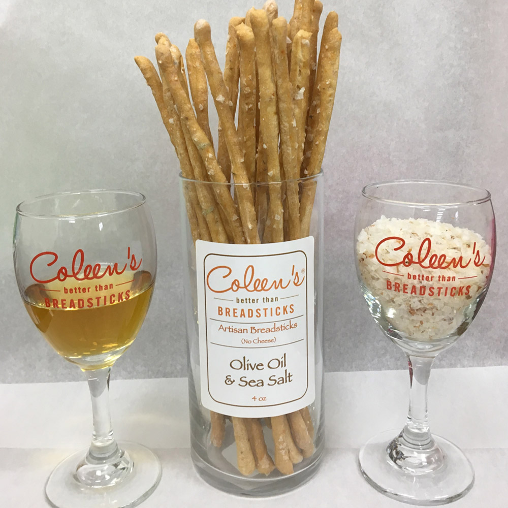 Glass container of Coleen's olive oil sea salt breadsticks next to two wine glasses
