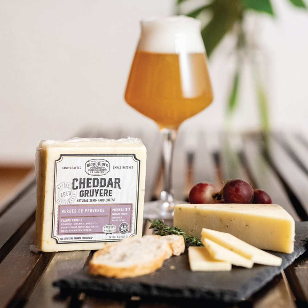 A block of Wood River Herbes de Provence Cheddar next to a cheese board and a glass of beer