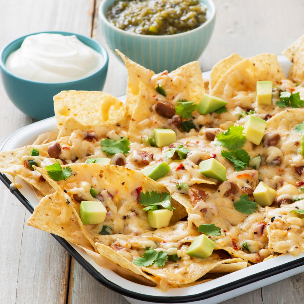 A plate of nachos next to a bowl of sour cream and a bowl of green salsa