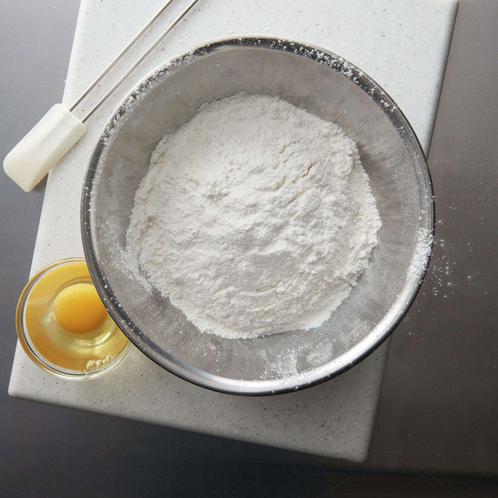 Cake flour in a bowl next to a bowl of eggs and spatula on top of a cutting board