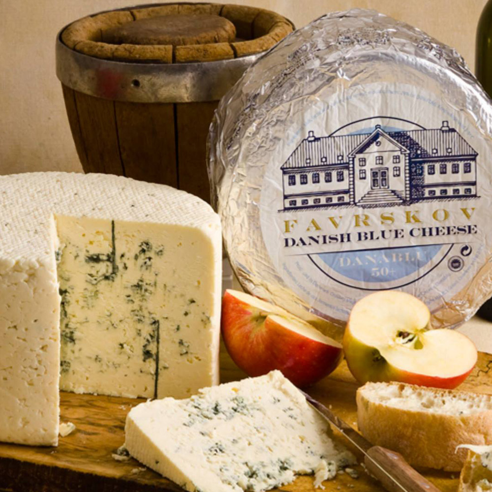 A wheel of Favrskov Danish blue cheese on a wooden board with apples and a bucket