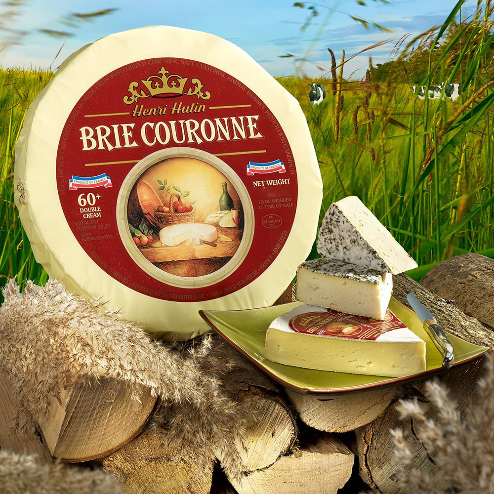 Wheel of Couronne brie on a pile of wood with grass in the background next to a plate with a wedge of brie