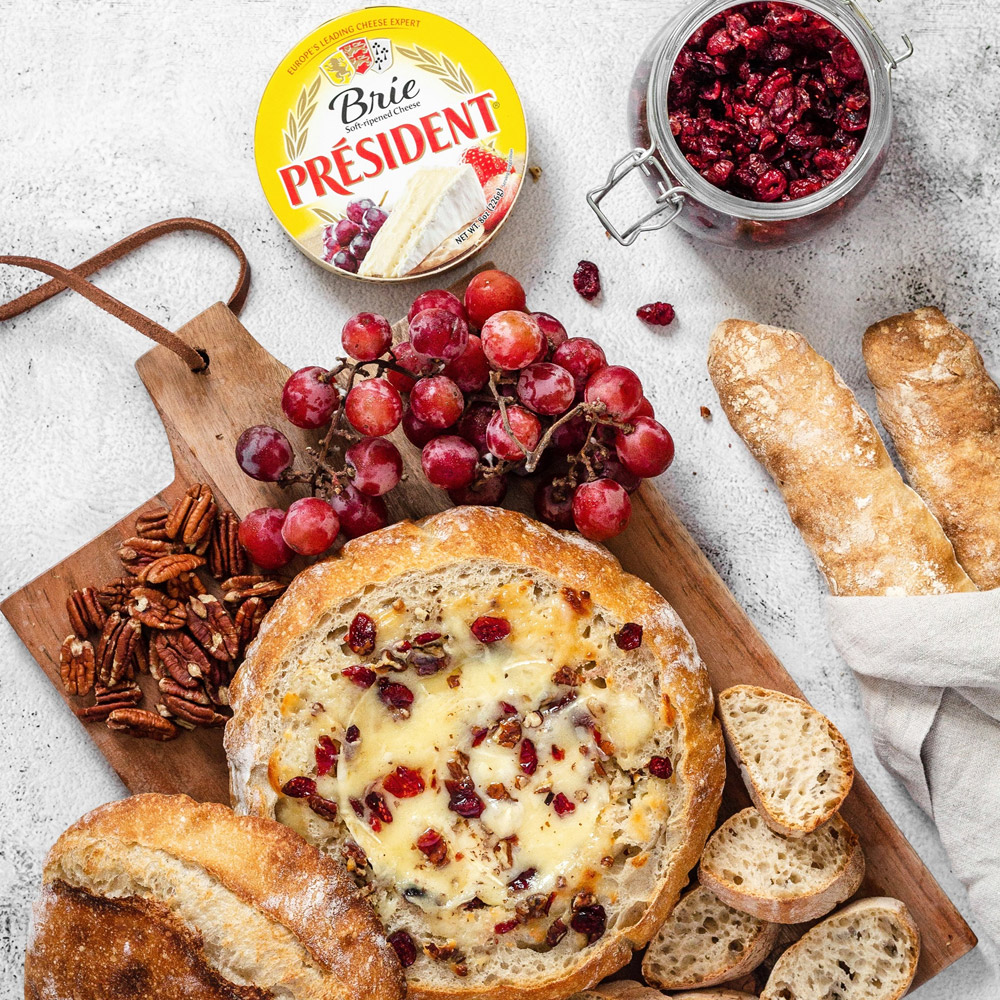 A package of President brie next to a board with a baked brie and some bread and a jar of cranberries