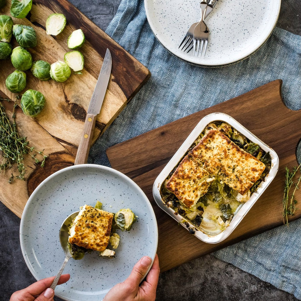 A tin on marinated grilling cheese on a wood board next to a person holding a plate with cheese and brussels sprouts