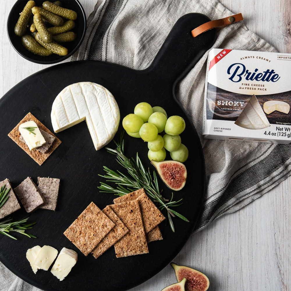 A package of Briette Smoky next to a cheese board with crackers, pate and fruit