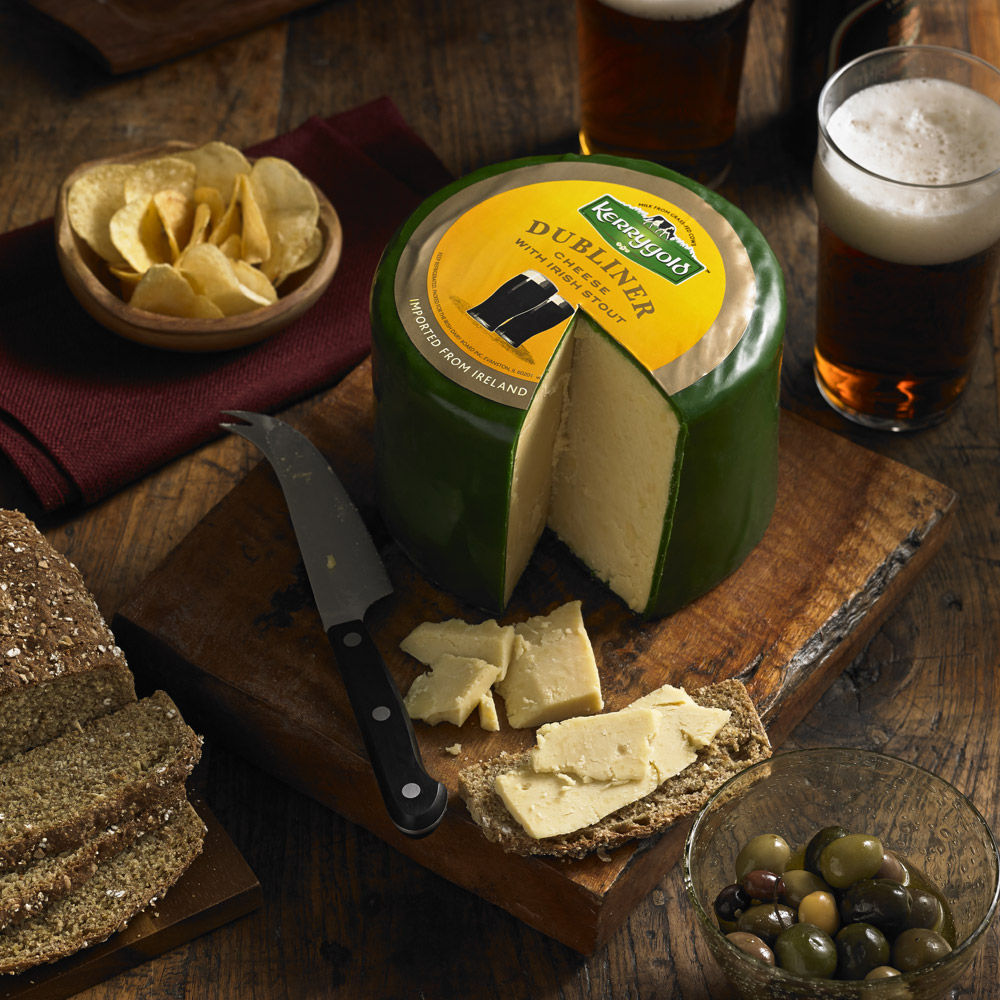 Kerrygold Dubliner cheese with Irish stout on a cutting board next to a bowl of potato chips and a bowl of olives