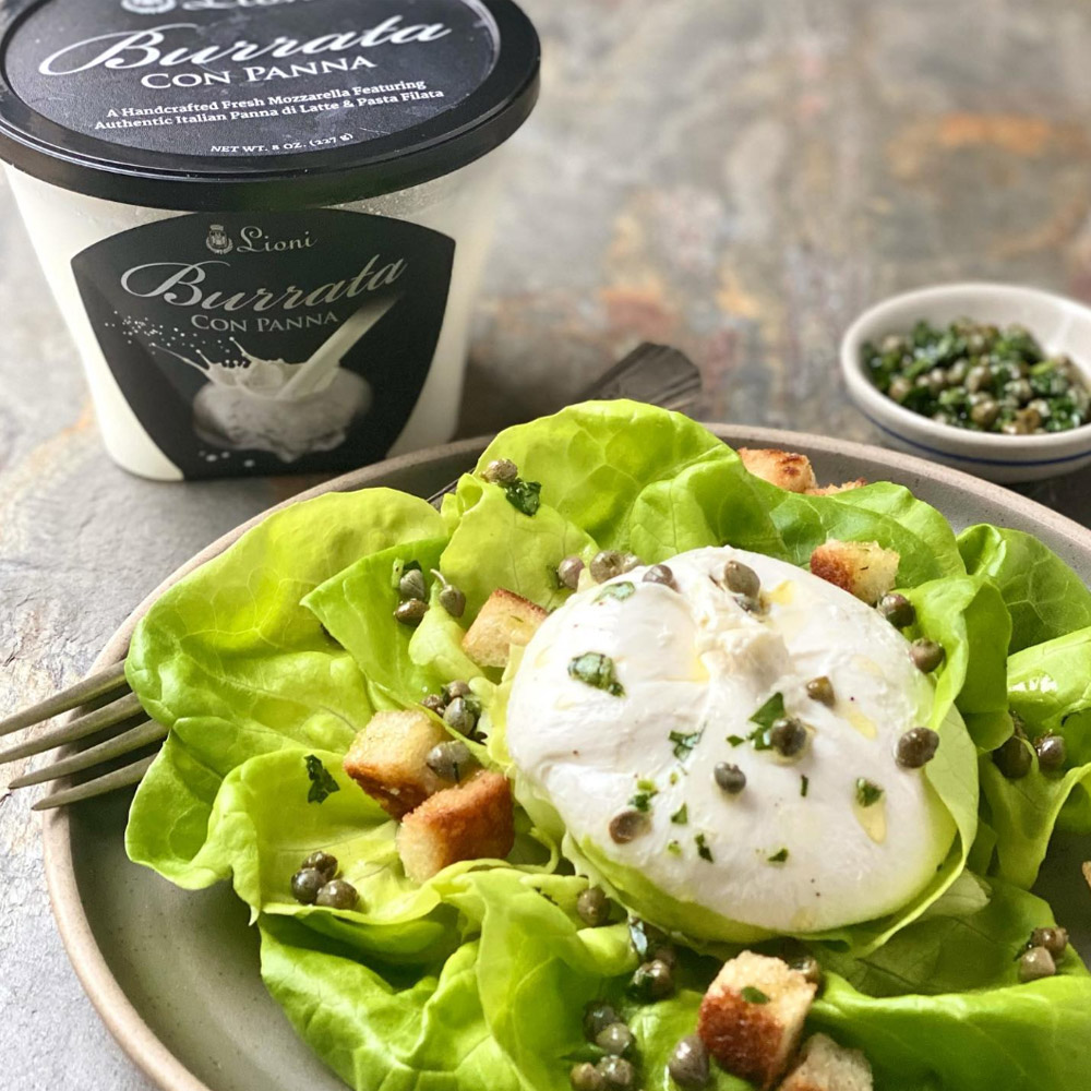 A container of Lioni Latticini Burrata behind a plate with a piece of burrata on top of lettuce topped with capers and croutons