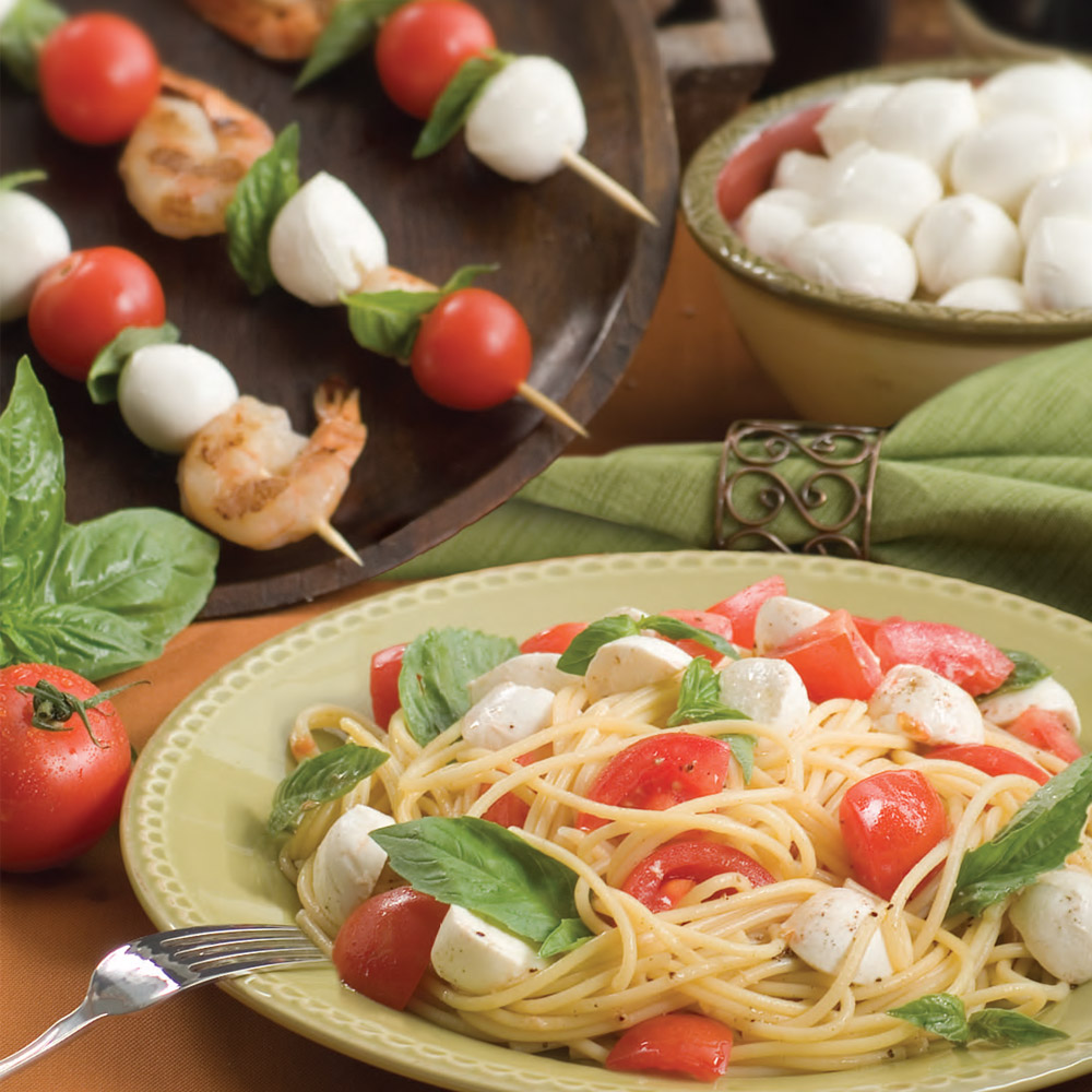 A bowl of spaghetti filled with tomatoes, basil and mozzarella in front of skewer with shrimp and mozzarella and a bowl of ciliegine