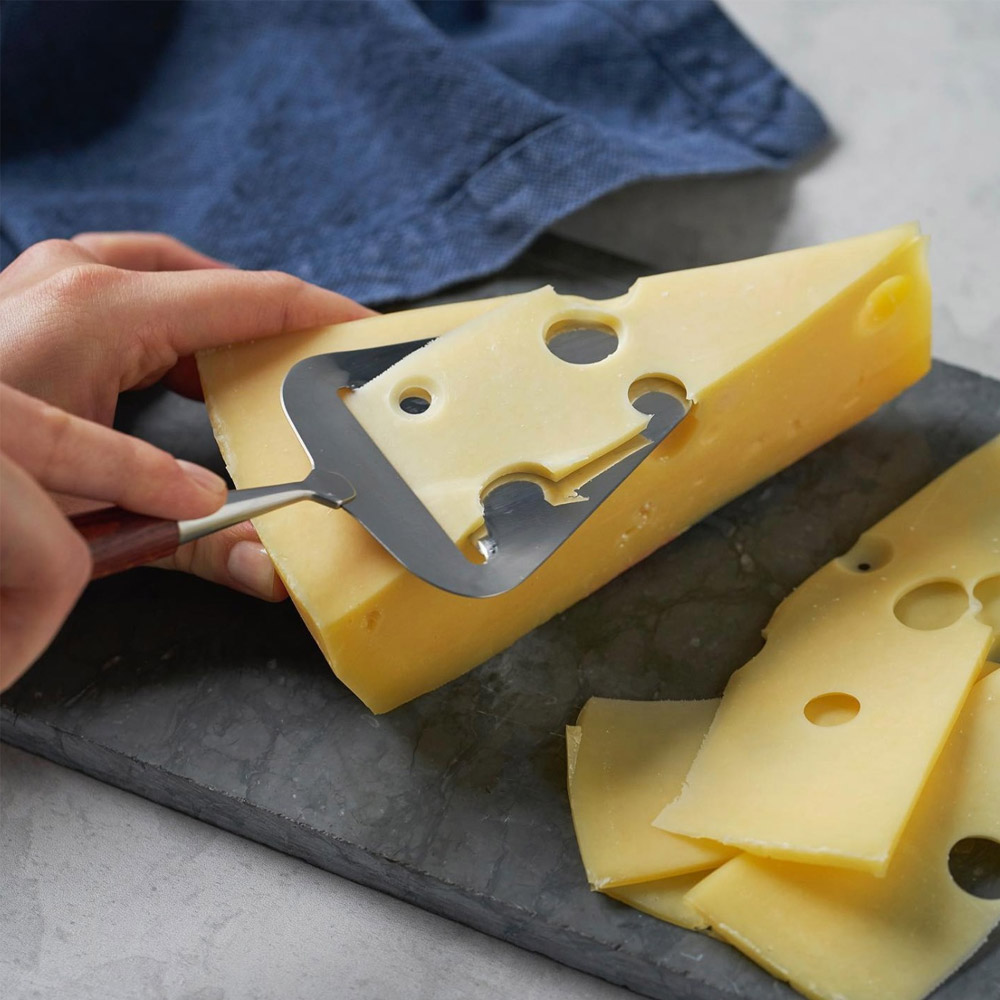 A wedge of swiss cheese on a slate board with a person slicing off a piece with a cheese cutter