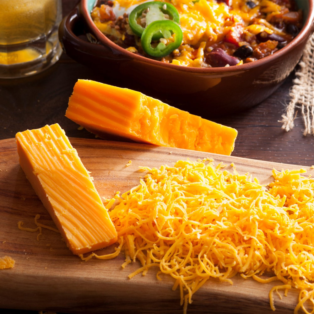 A grated block of cheese next to a pile of grated cheese in front of a bowl of chili with cheese on top
