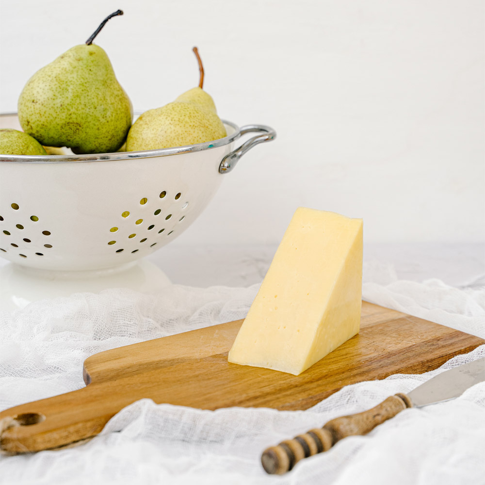 A wedge of white cheddar on a wood board in front of a strainer filled with green pears