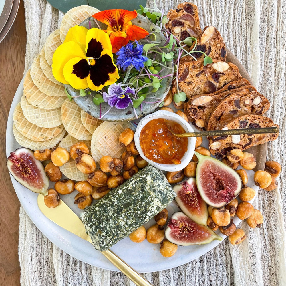 A cheese board with a herb goat cheese log, snacks and flowers