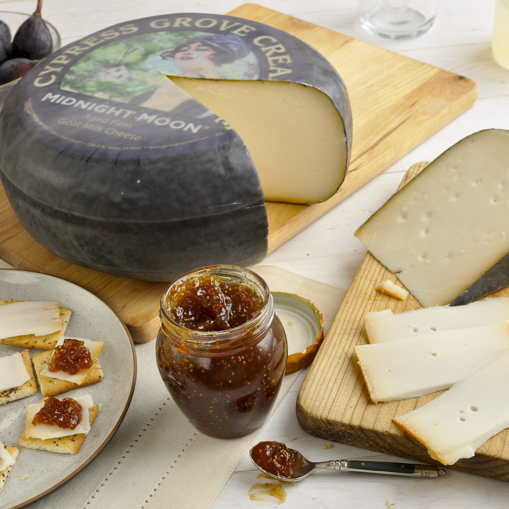 A wheel of Cypress Grove Midnight Moon cheese next to a wedge of cheese with an open jar of fig jam