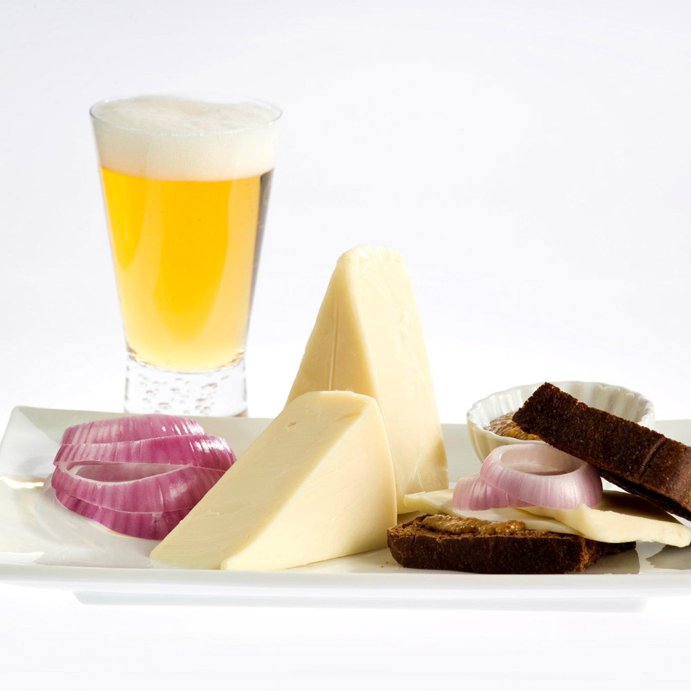 Two wedges of Benedictine cheese on a white plate with onions, brown bread and a bowl of mustard next to a glass of cold beer
