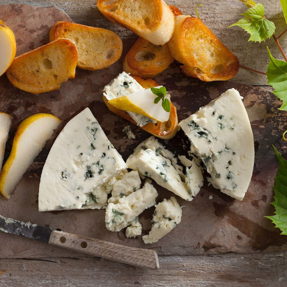 Two wedges of blue cheese on a wood surface with slices of pear and toasted bread
