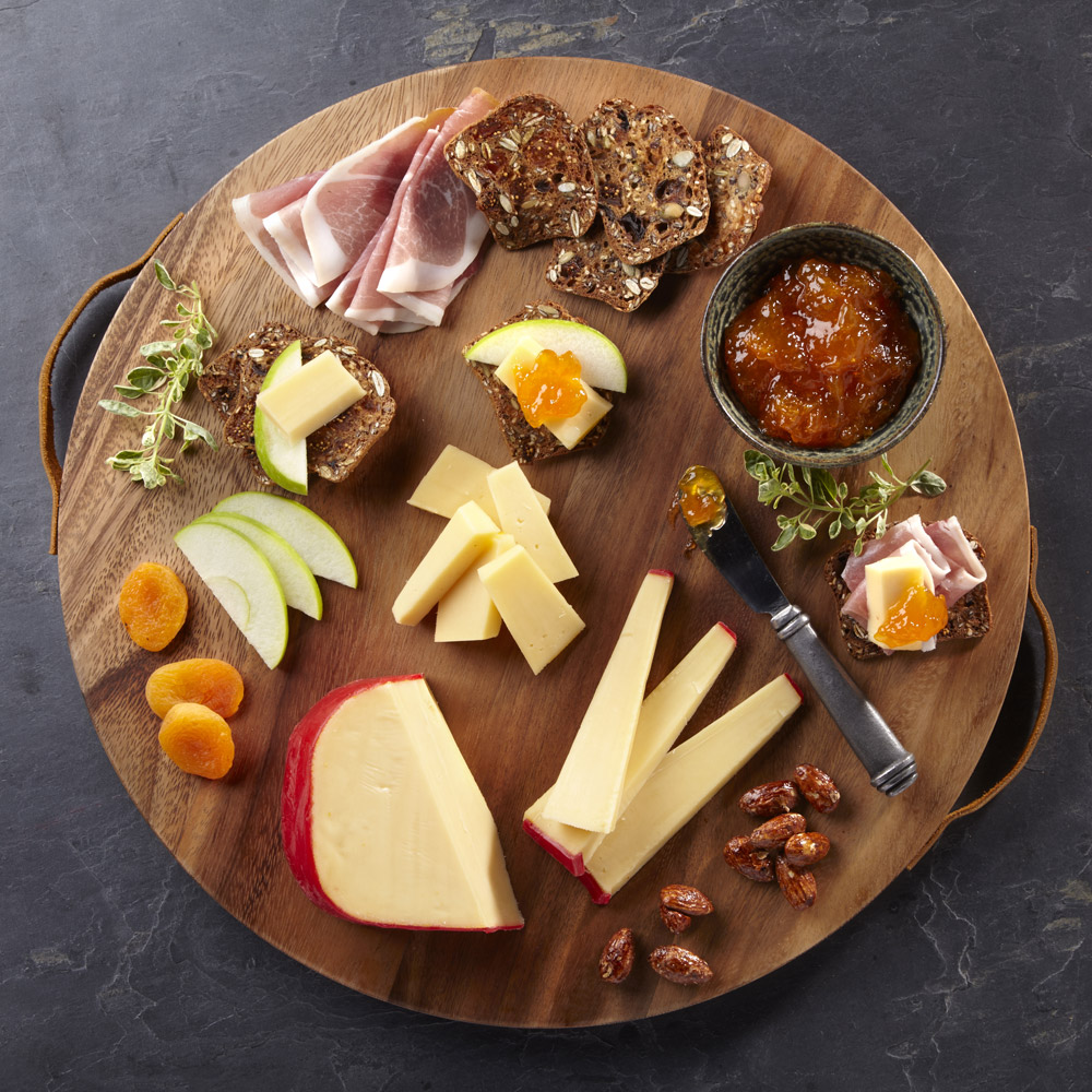 A wedge of gouda on a wood board with accompaniments