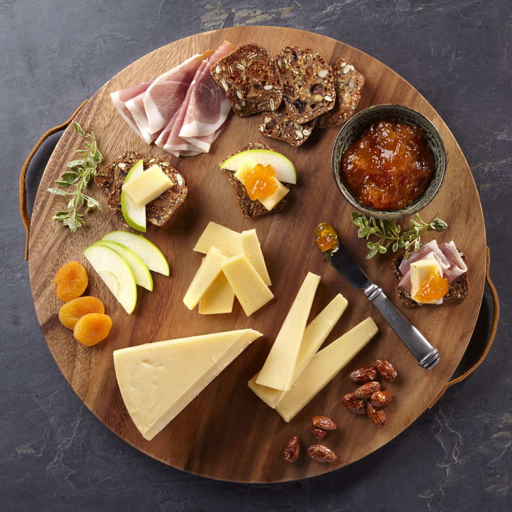 A wedgge e of gouda on a wood board with accompaniments