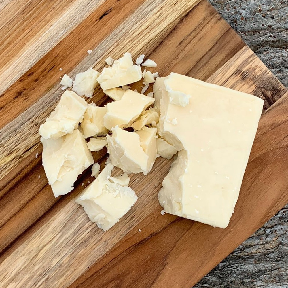 A piece of white cheddar next to white cheddar crumbles on a wood board