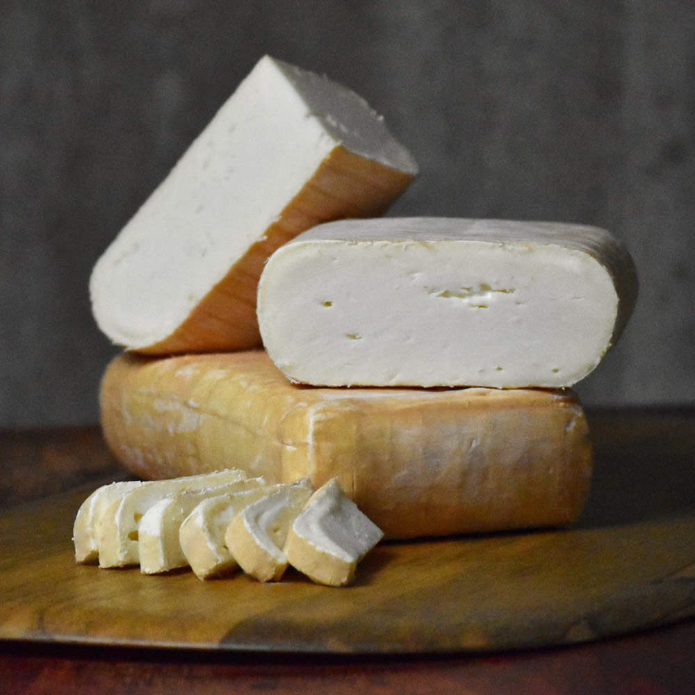 A brick of Eligo cheese under a second brick cut in half with slices of Eligo on a cheese board