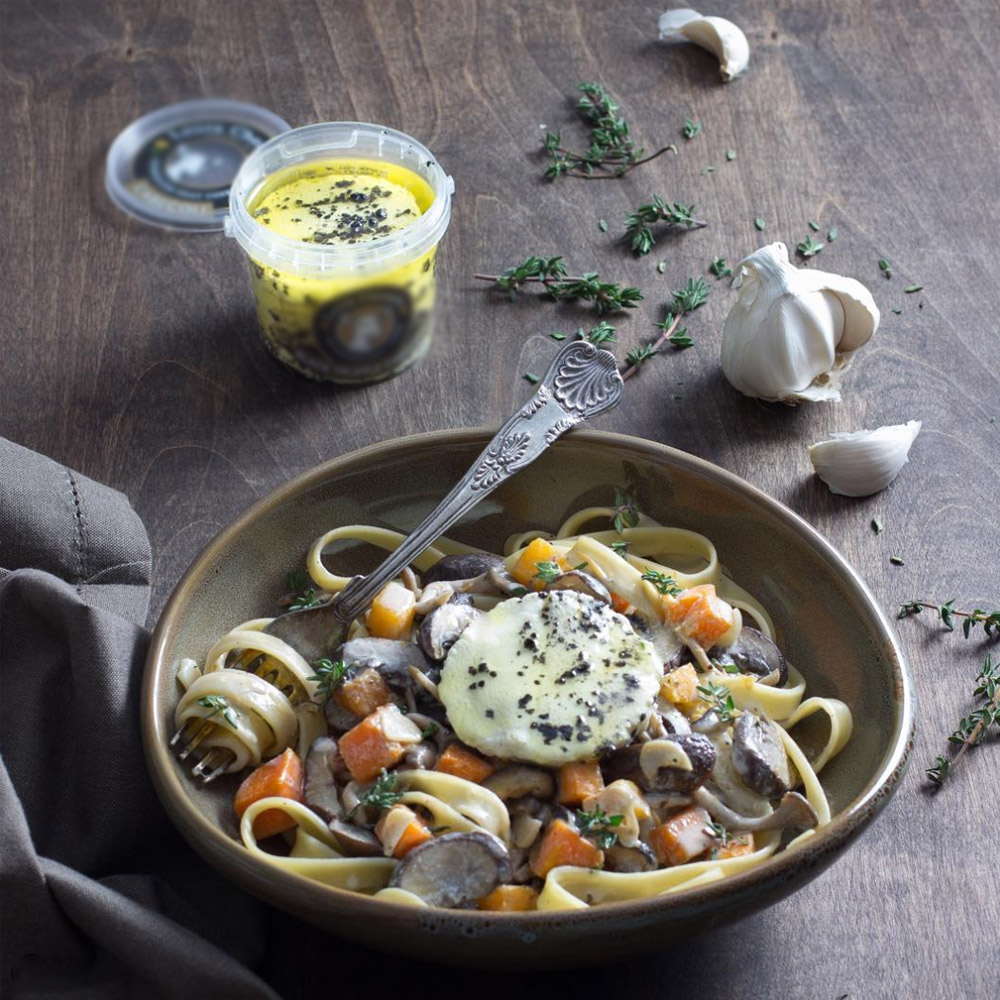 A bowl of fettuccine pasta topped with vegetables and a slice of marinated goat cheese in front of a container of marinated goat cheese
