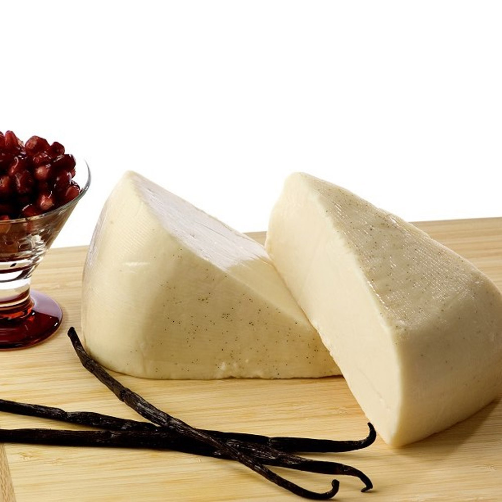 Two wedges of Carr Valley Sweet Vanilla Cardona cheese on a wood board with some vanilla beans and a glass cup of pomegranate seeds