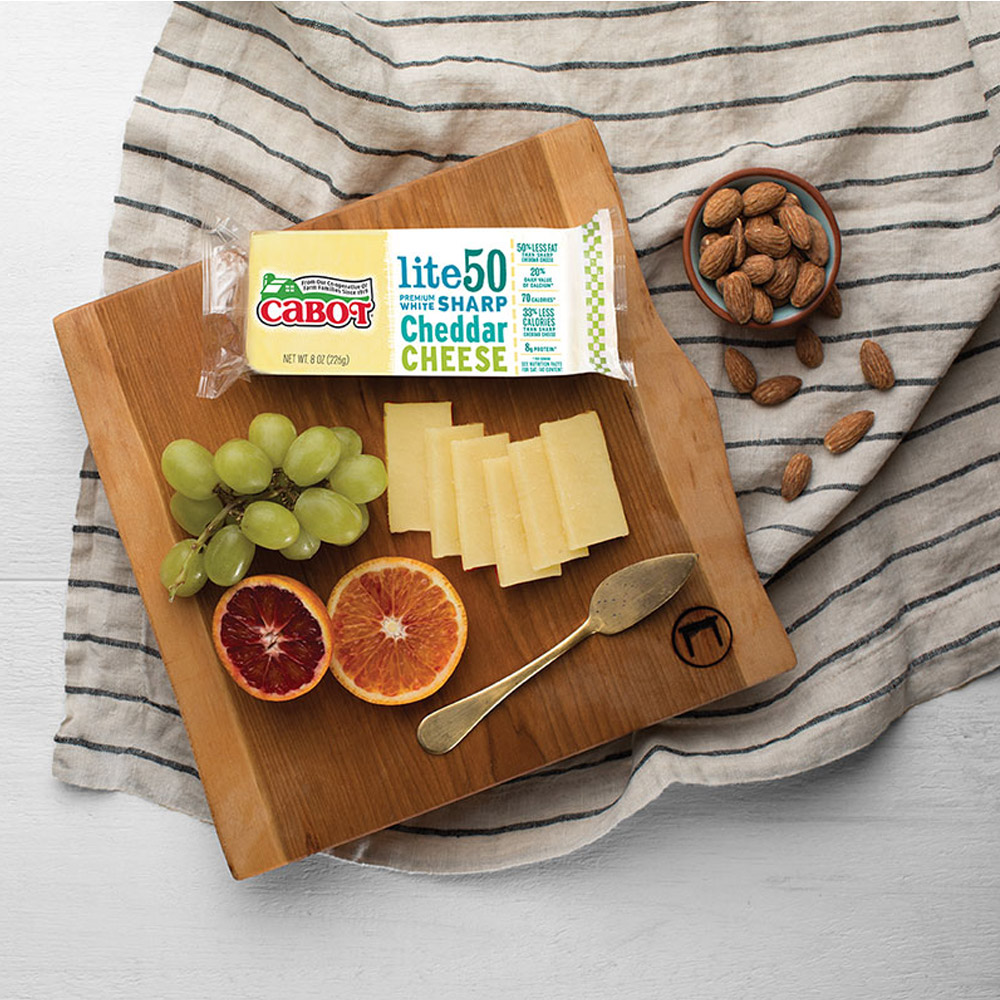 A bar of Cabot Lite50 Sharp Cheddar on a board with fruit and a cheese knife next to a cup of almonds