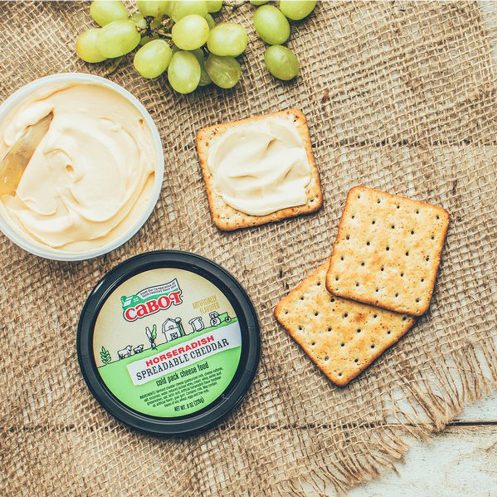 Open container of Cabot Horseradish Cheddar Spread next to grapes and crackers on a burlap background