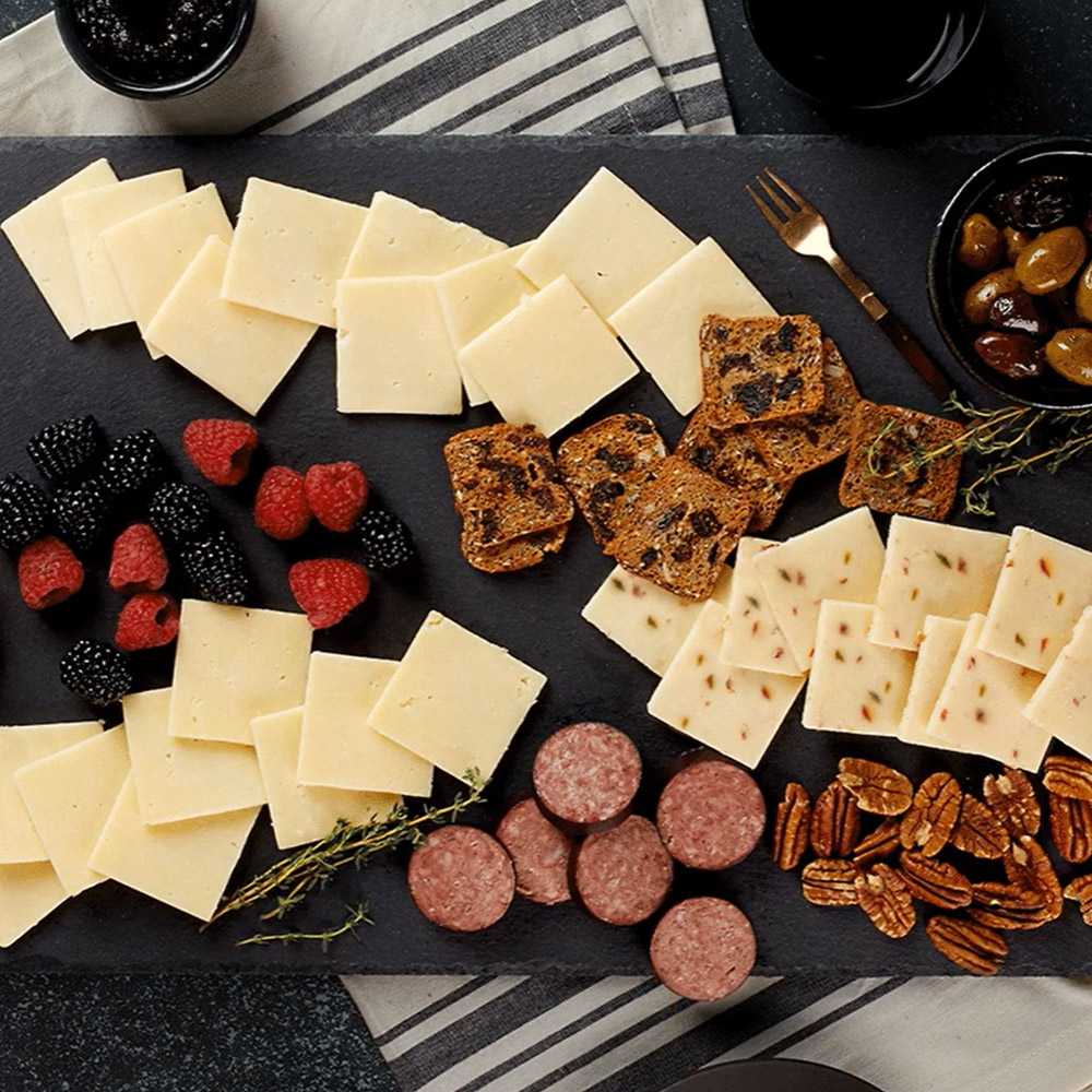 A slate board with slices of white cheddar cheese, berries, salami, crackers and herbs next to a bowl of olives
