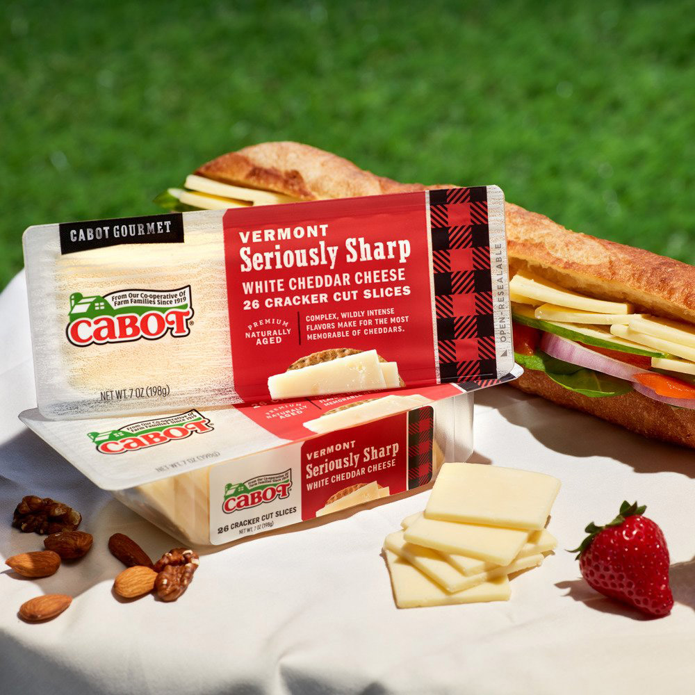 Two packages of Cabot seriously sharp white cheddar cracker cuts on a wood board with fruit and nuts beside and a sandwich in the background