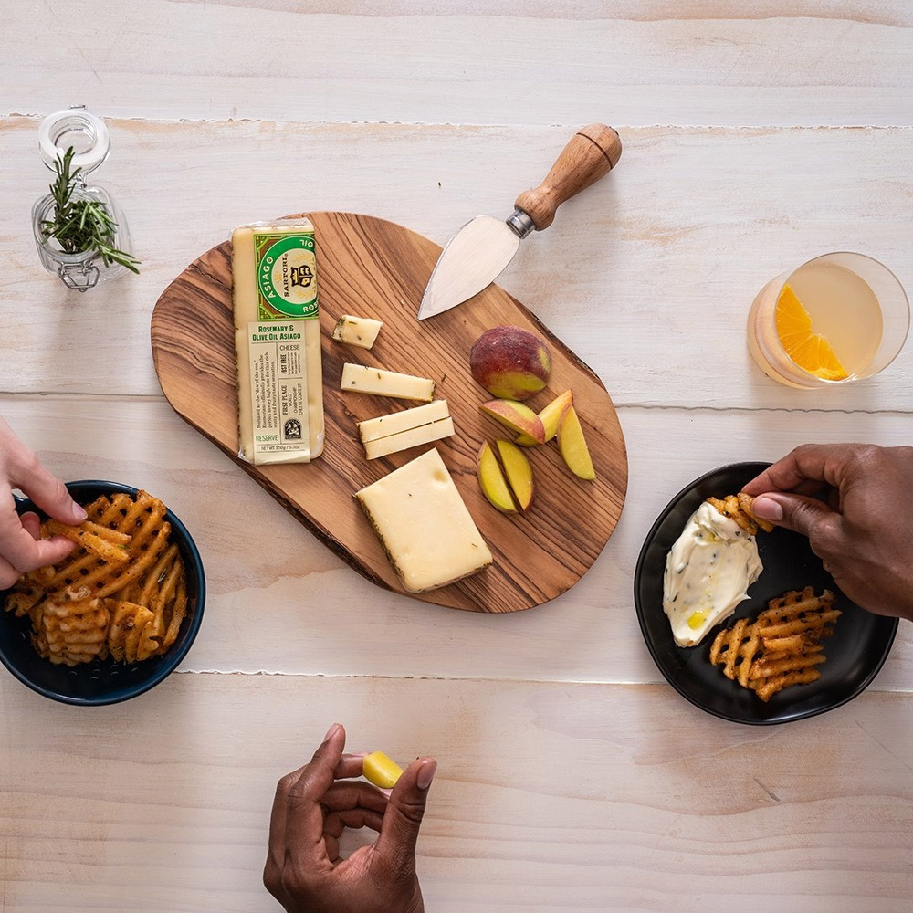 A cheese board with a wedge of asiago cheese next to hands eating waffle fries and dip