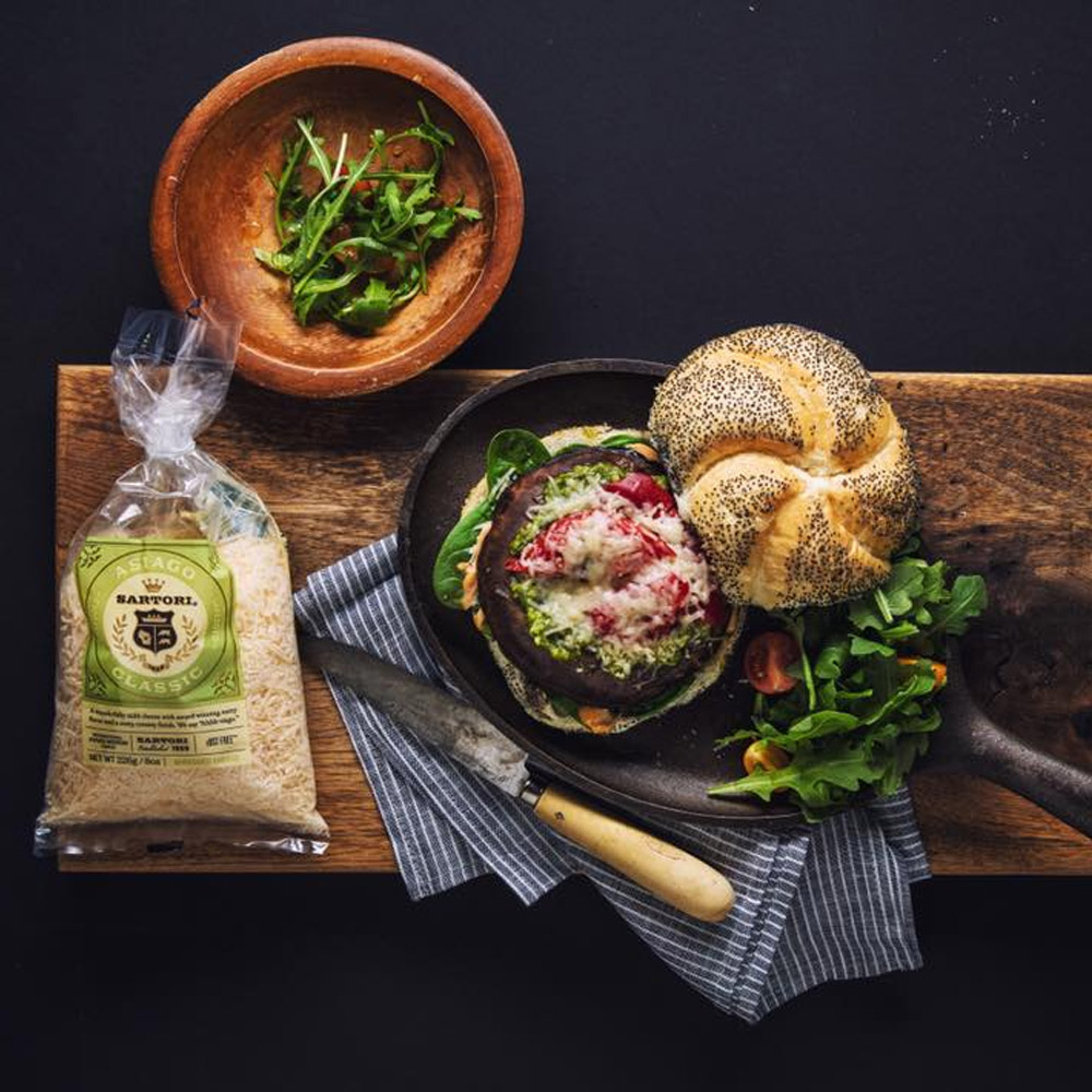 A bag of Sartori asiago cheese next to a burger on a cast iron skillet and a bowl of greens