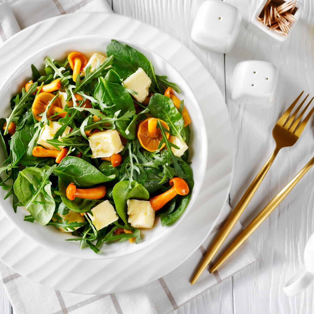 Arugula, spinach, honey mushrooms, and cubed cheddar cheese salad in a white bowl