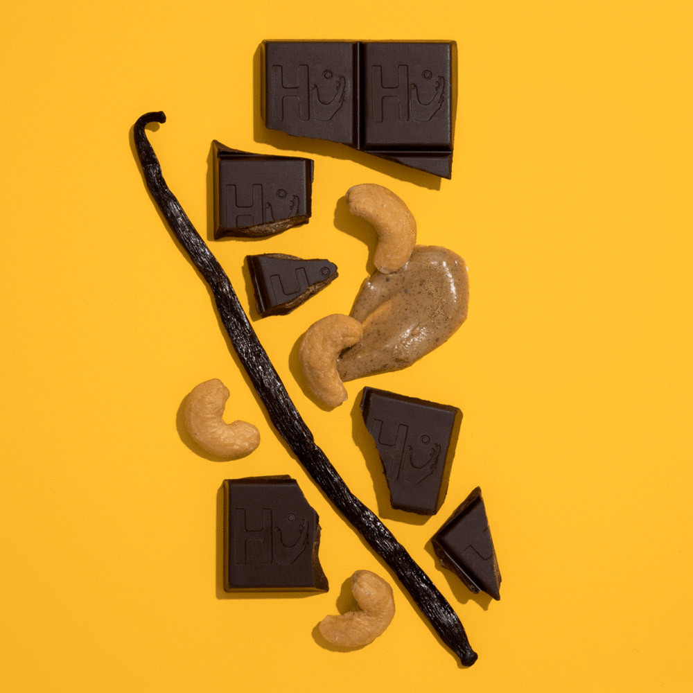 A broken Hu chocolate bar next to a vanilla bean and cashews on a yellow background