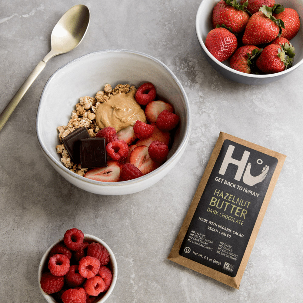 A Hu Hazelnut Butter Organic Dark Chocolate Bar next to two bowls of berries and one bowl with berries and granola with hazelnut butter