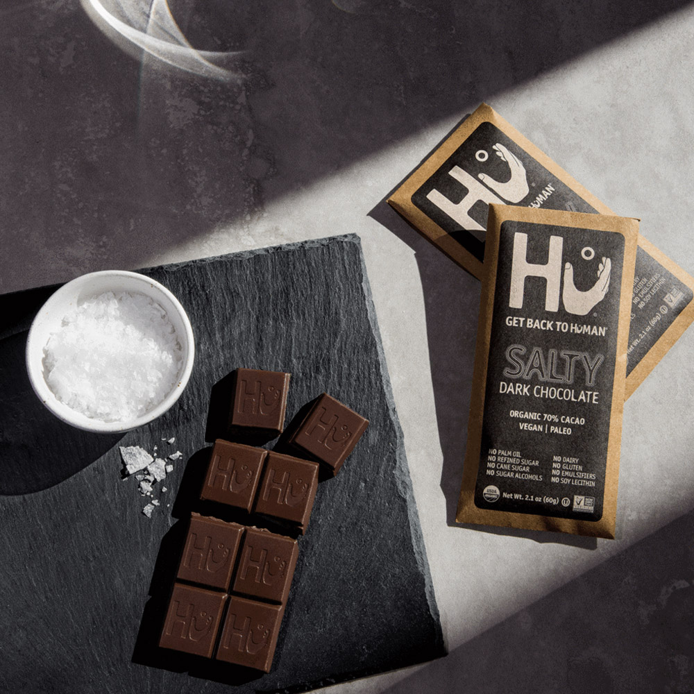 Two Hu Salty Organic Dark Chocolate Bars next to an unwrapped bar that is next to a bowl of rock salt