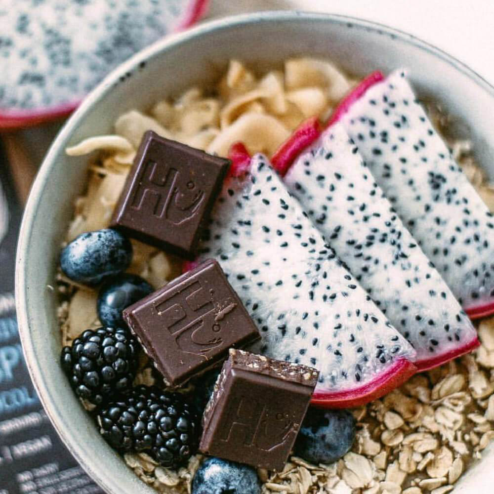 Pieces of a Hu Vanilla Crunch Organic Dark Chocolate Bar in a bowl with fruit and oats