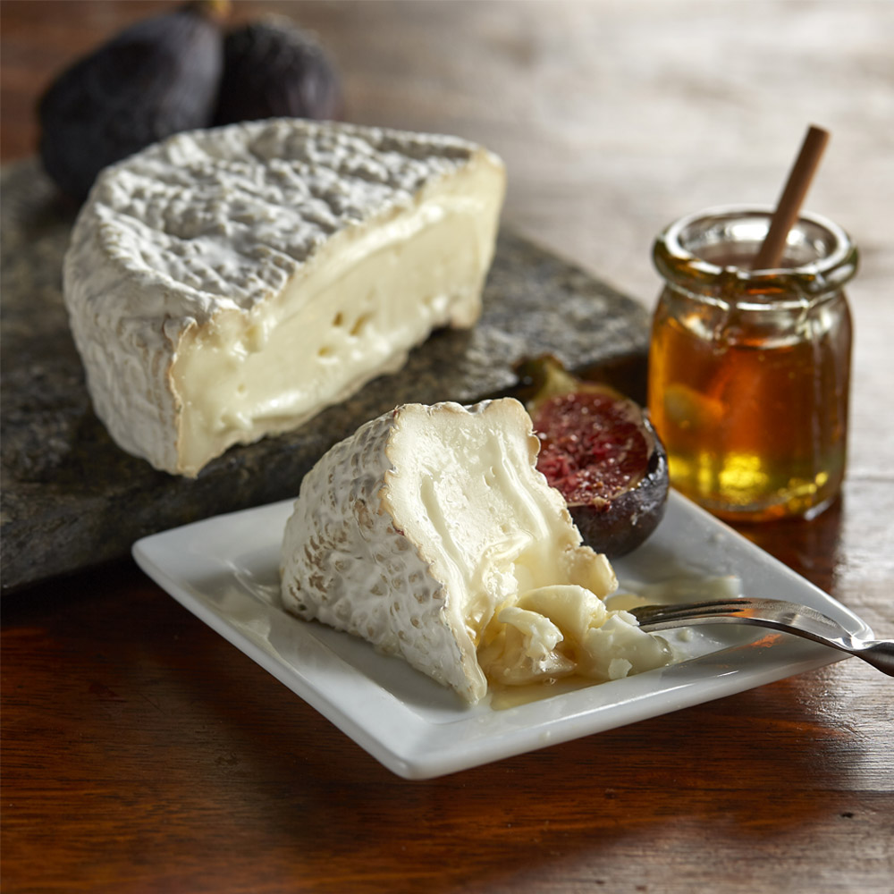 A piece of Merry goat Round cheese on a board next to a wedge of cheese on a plate with honey and a fig
