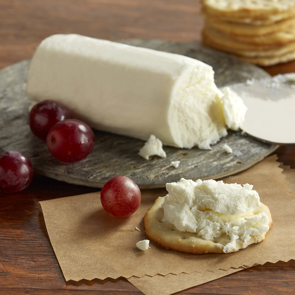 An unwrapped log on a board behind a cracker topped with goat cheese and some grapes