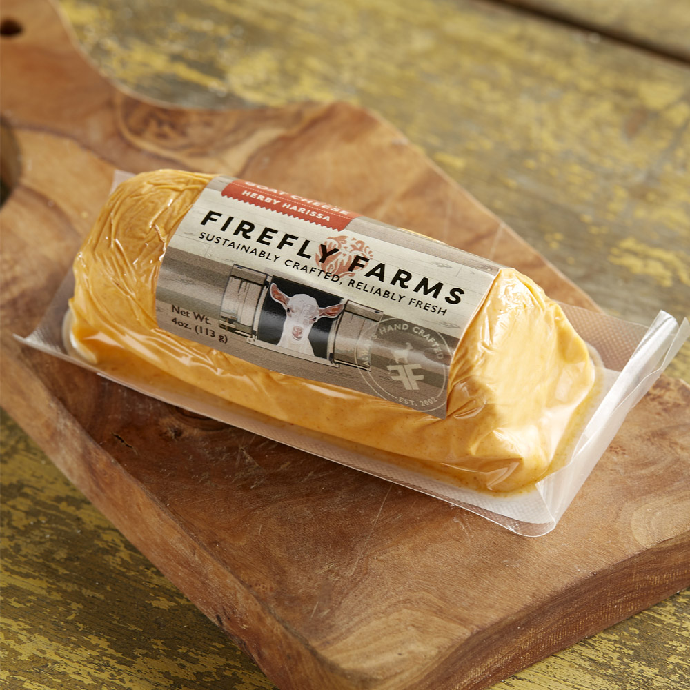 A log of Firefly Farms Herby Harissa fresh goat cheese in its packaging on a wood board