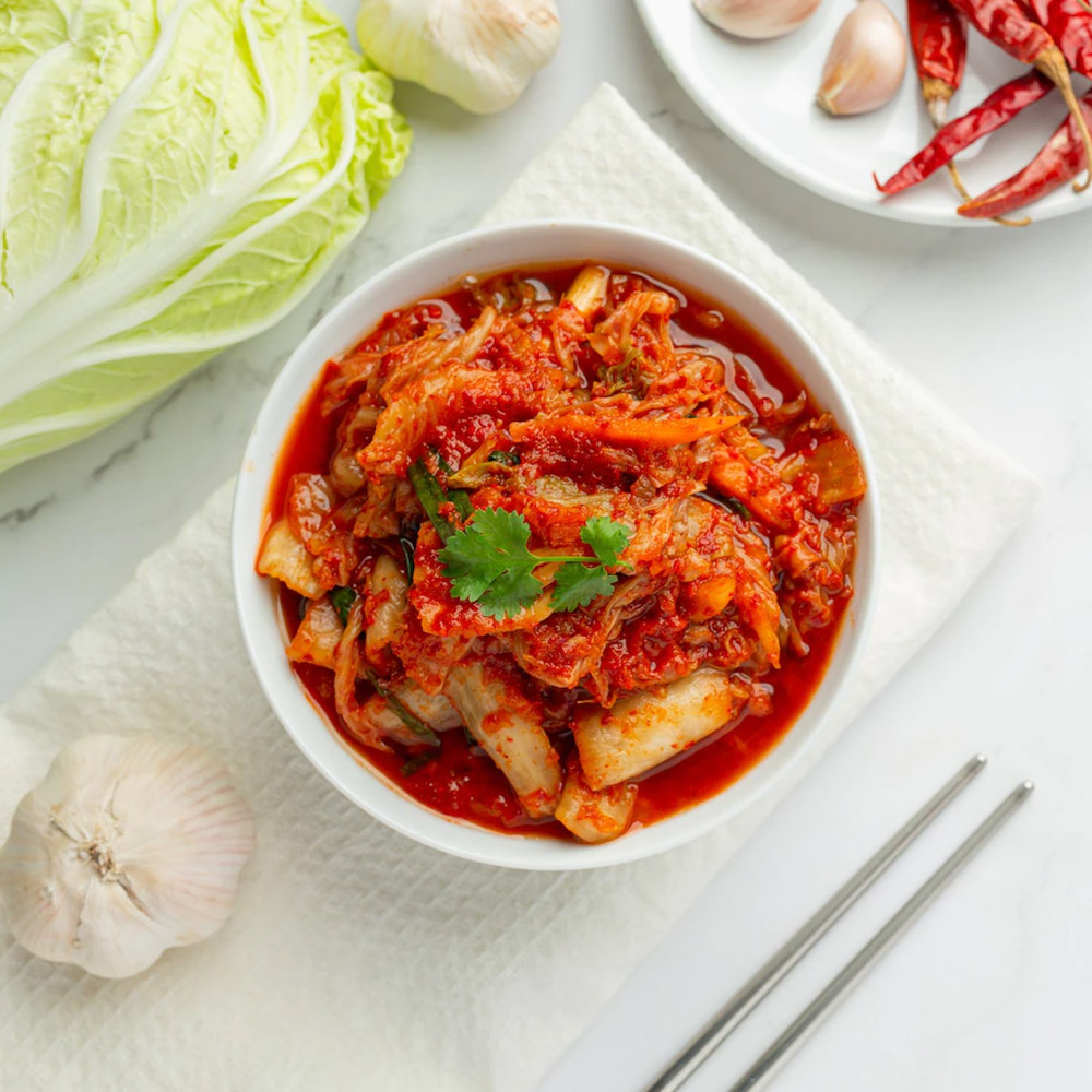 A bowl of kimchi next to garlic and cabbage and a plate of peppers