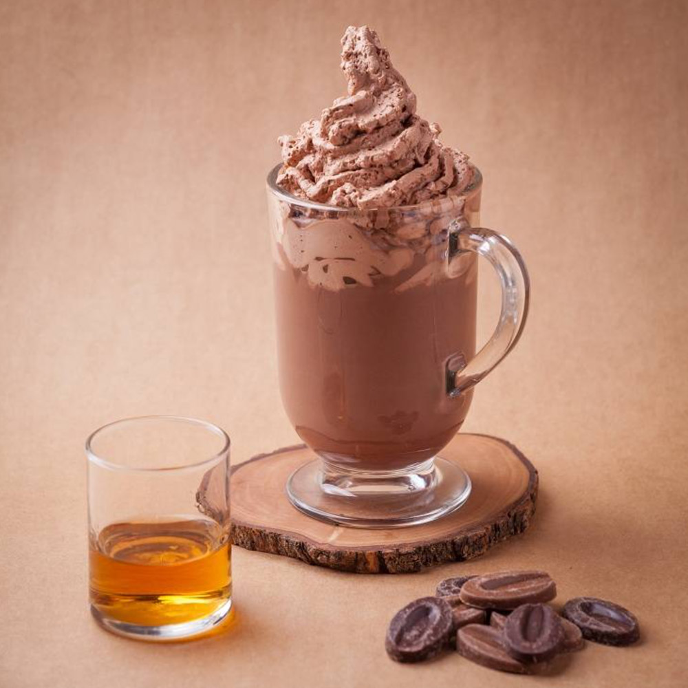 A glass of hot chocolate on a wood plank next to a glass of whiskey and a pile of chocolate feves
