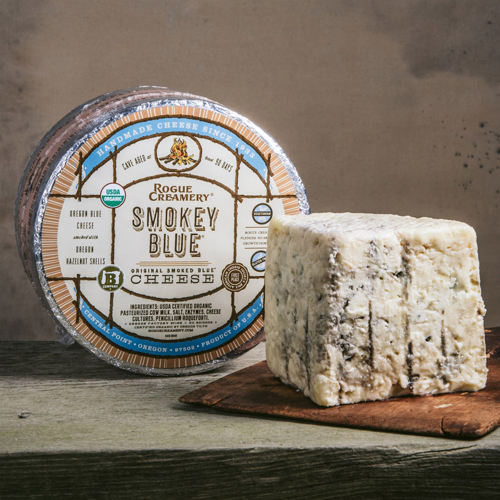 Rogue Creamery organic Smokey blue cheese wheel next to a wood board with a wedge of blue cheese