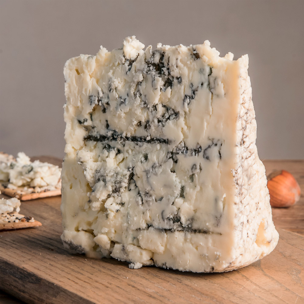 A wedge of blue cheese on a wood board next to a nut and a cracker topped with blue cheese