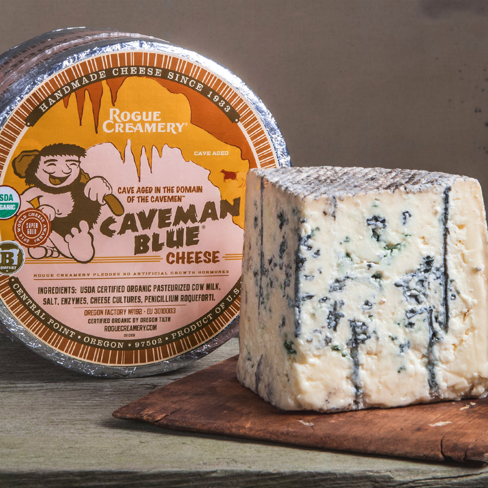 Rogue Creamery organic Caveman blue cheese wheel next to a wood board with a wedge of blue cheese