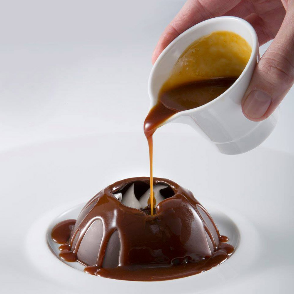 An upside down chocolate grand bowl on a plate with a person's hand pouring caramel on top