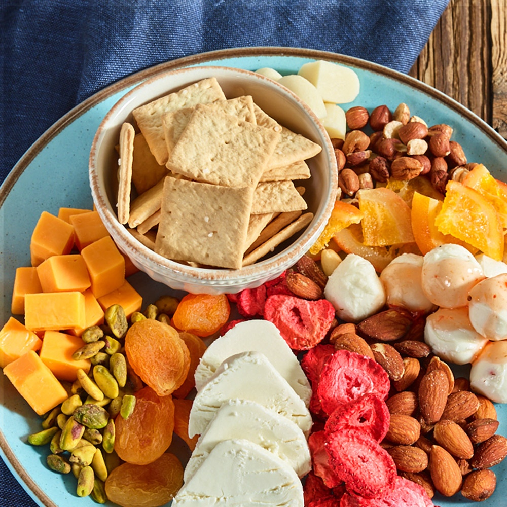 A bowl of crackers surrounded by cheese and fruit and nuts