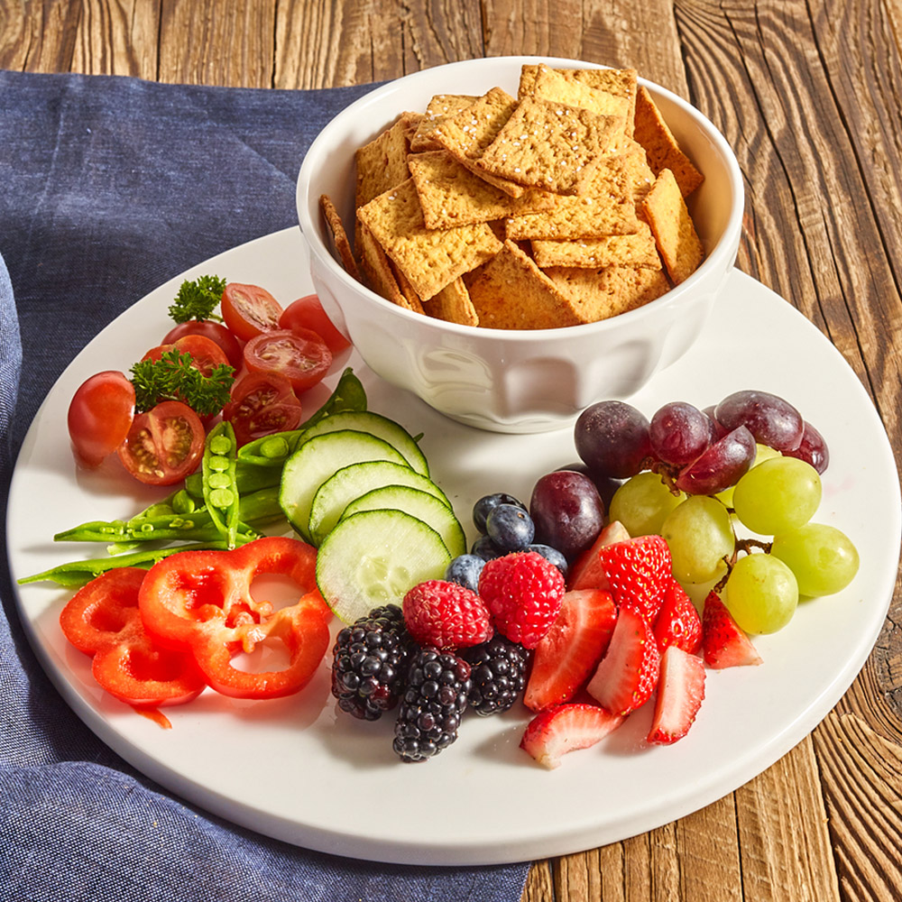 A bowl of crackers on a plate surrounded by fruits and vegetables