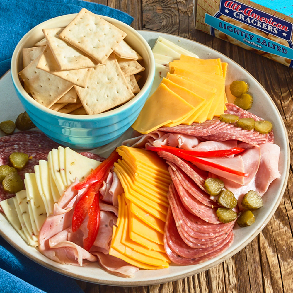 A bowl of crackers on a plate surrounded by cheese and salami