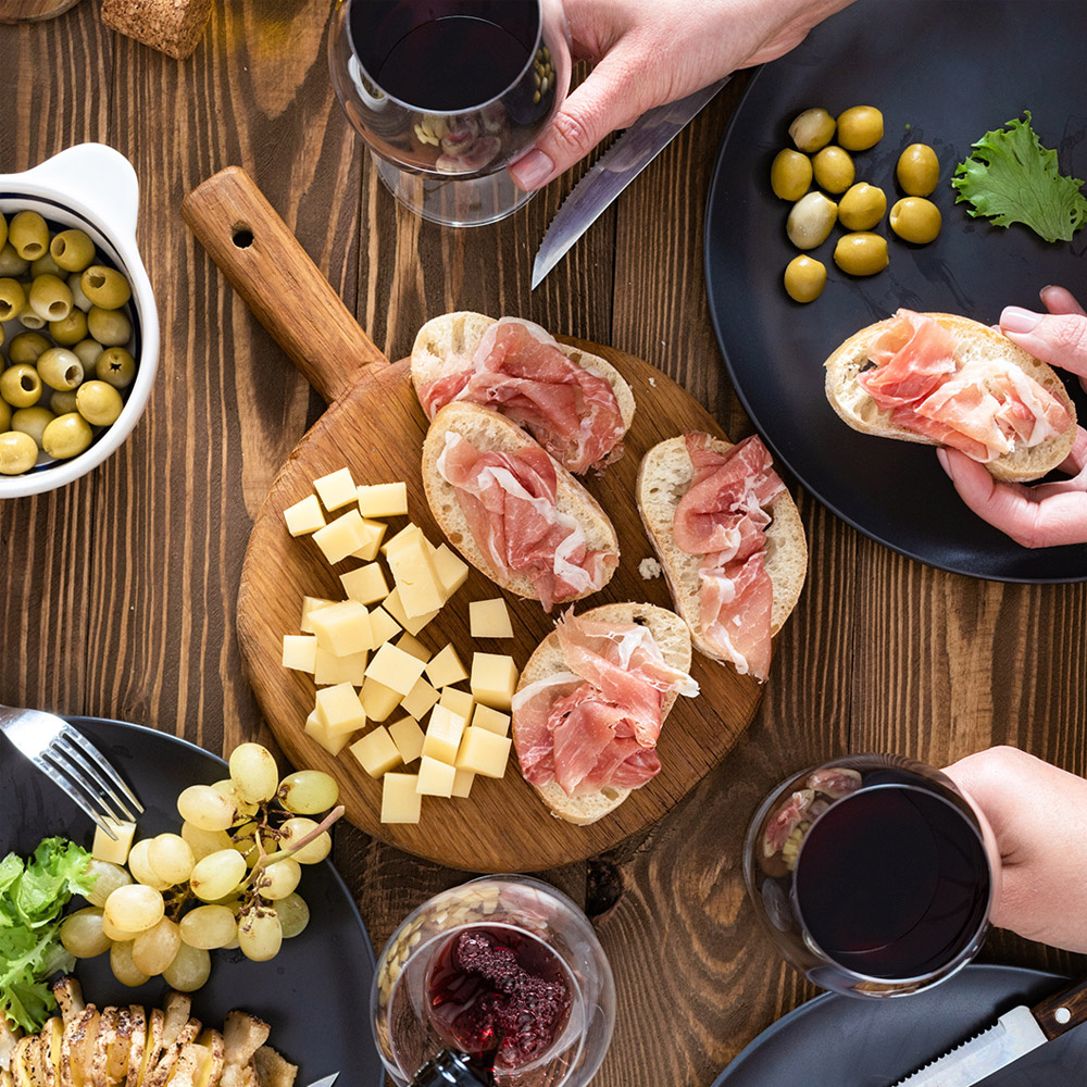 A board with bread that is topped with prosciutto surrounded by wine glasses and plates with fruit and other snacks