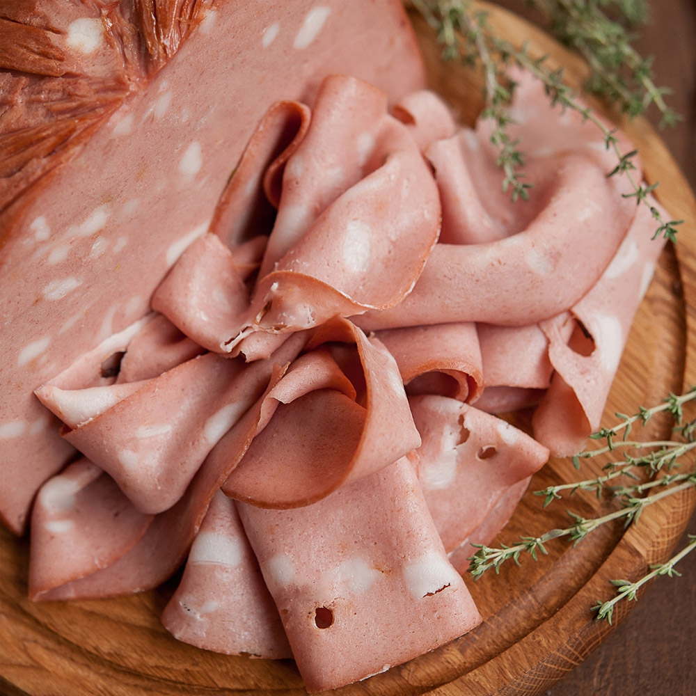 A chunk of mortadella on a wood board next to slices of mortadella and herbs